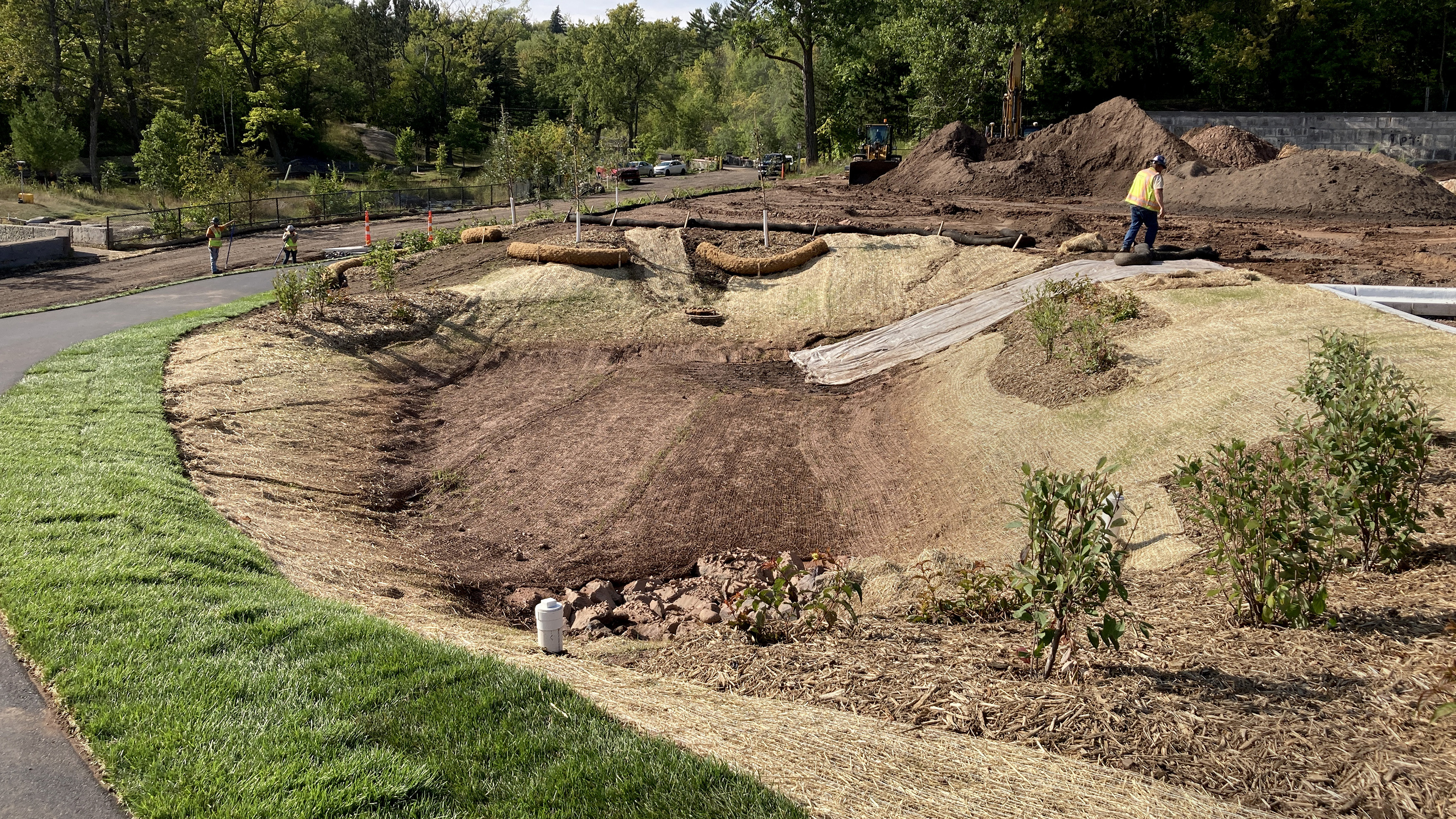 Tan erosion control material lines a basin ringed by mulched shrubs and a paved trail