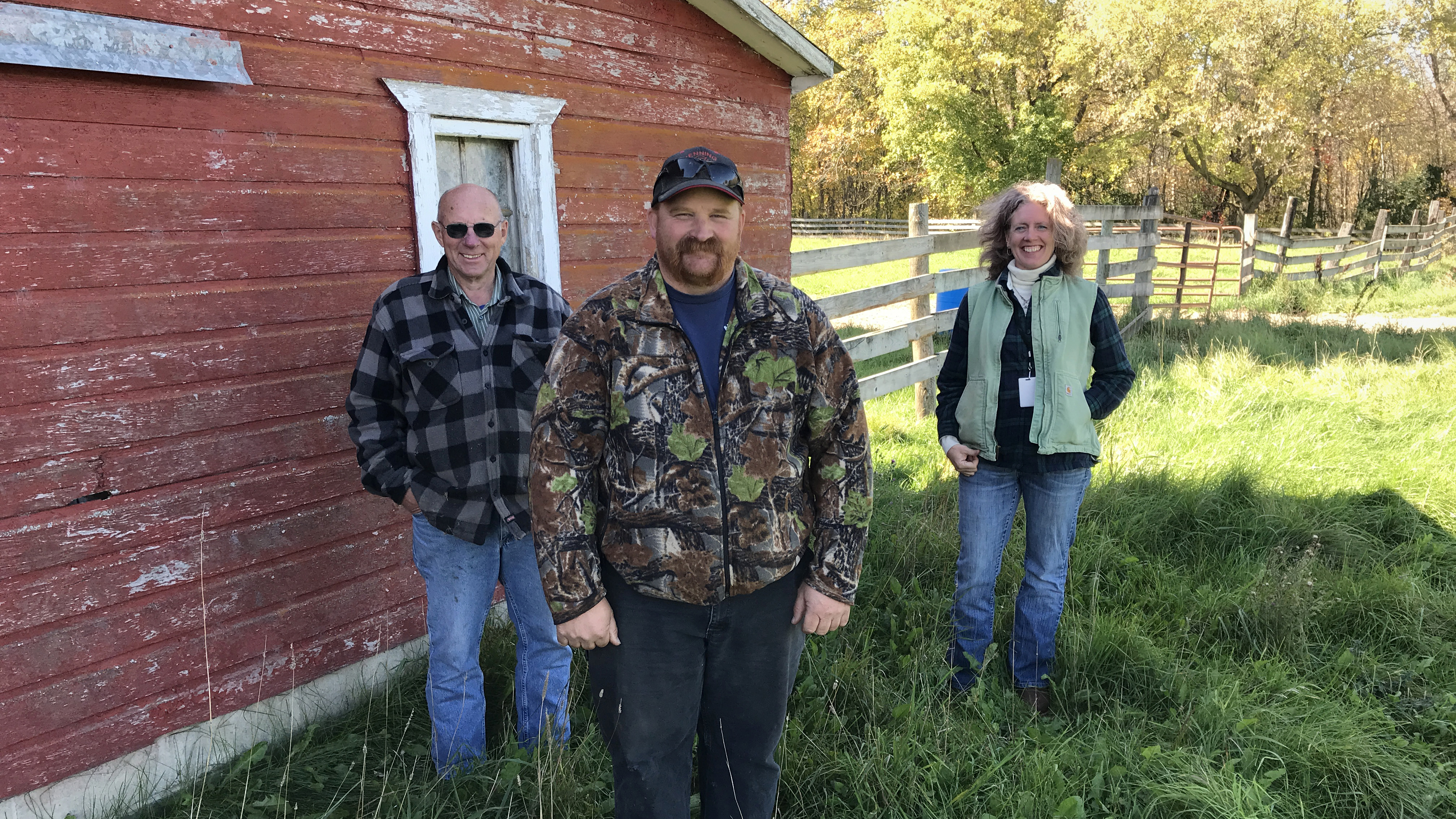 three people stand along the side of a red barn. a wooden fence is in the background