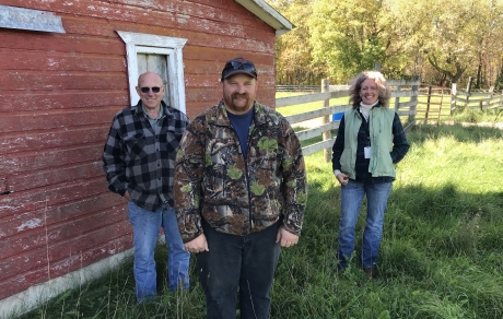 three people stand along the side of a red barn. a wooden fence is in the background